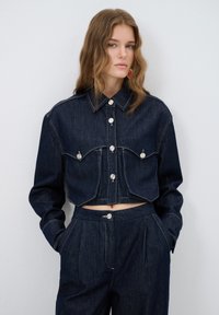 Tummansininen lyhyt takki, jossa on suuret taskuläpät ja hopeiset painonapit, yhdistettynä leveisiin housuihin, joissa on samanlainen nappi.