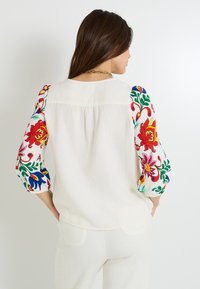 Blusa blanca con bordado floral colorido en las mangas. Tejido suave, corte holgado, detalle fruncido en la parte posterior y puños elásticos.