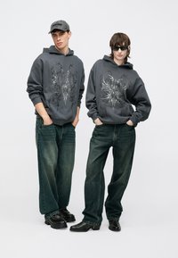 Tmavohnedé mikiny s grafickou potlačou, voľné modré rifle a masívne čierne topánky. Doplnky zahŕňajú sivú šiltovku a slnečné okuliare.