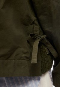 Κοντινό μιας σκουροπράσινης ζακέτας δεμένης με υφασμάτινο φιόγκο πάνω από ένα υφασμάτινο πλεκτό ένδυμα και ένα μπλε ριγέ πουκάμισο.