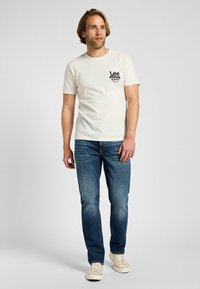 Camiseta de algodón crema con mangas cortas, que presenta un logo gráfico negro en el pecho, combinada con jeans de mezclilla azules y zapatillas blancas.