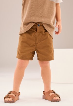 Småbarn iført brune shorts, en beige striktrøje og brune sandaler med spænder, stående på en ensfarvet lys baggrund.