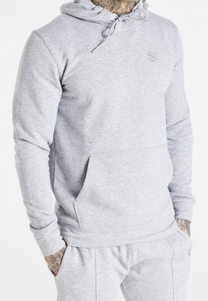 Mann trägt hellgrauen Hoodie und passende Jogginghose, Tattoos am Hals und an den Händen sichtbar, Hände in den Taschen.