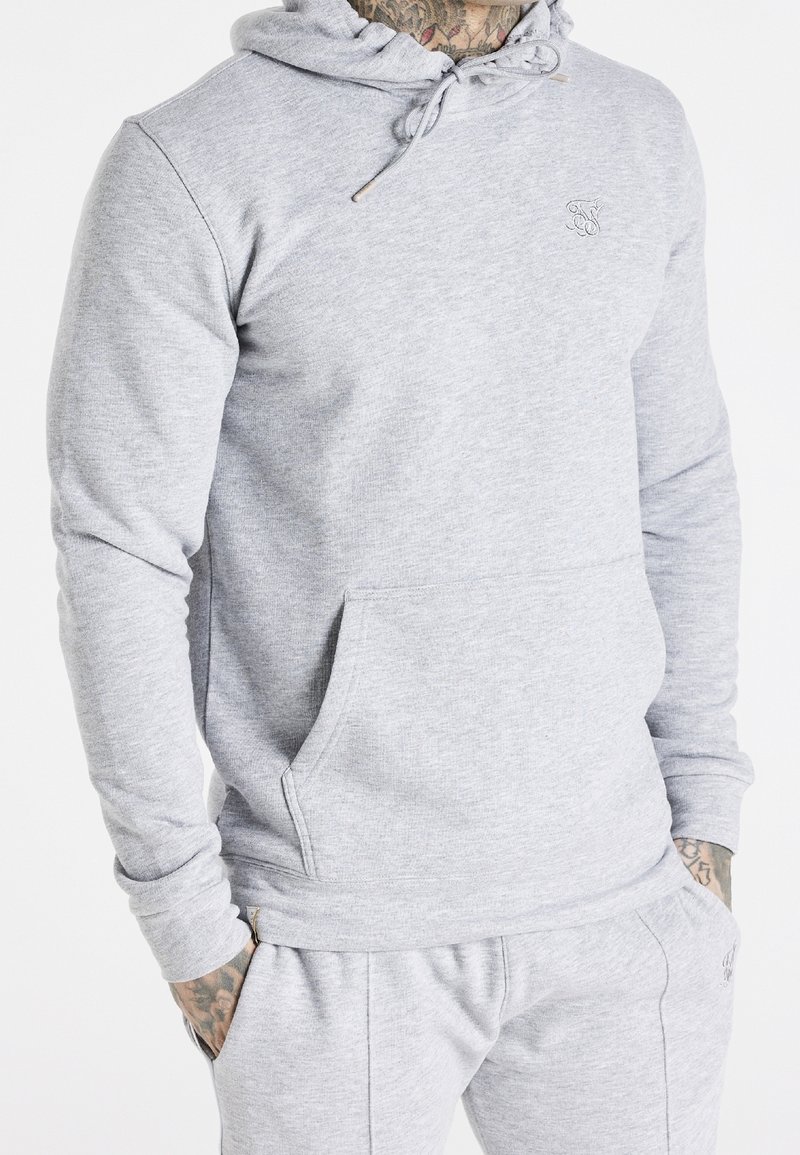 Homme portant un sweat à capuche gris clair et un pantalon de survêtement assorti, avec des tatouages visibles sur le cou et les mains, les mains dans les poches.