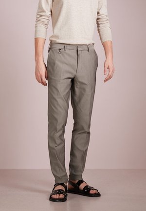 Homme portant un pull beige, un pantalon gris sur mesure avec passant de ceinture et détail anneau, et des sandales noires cloutées, debout sur un sol clair.