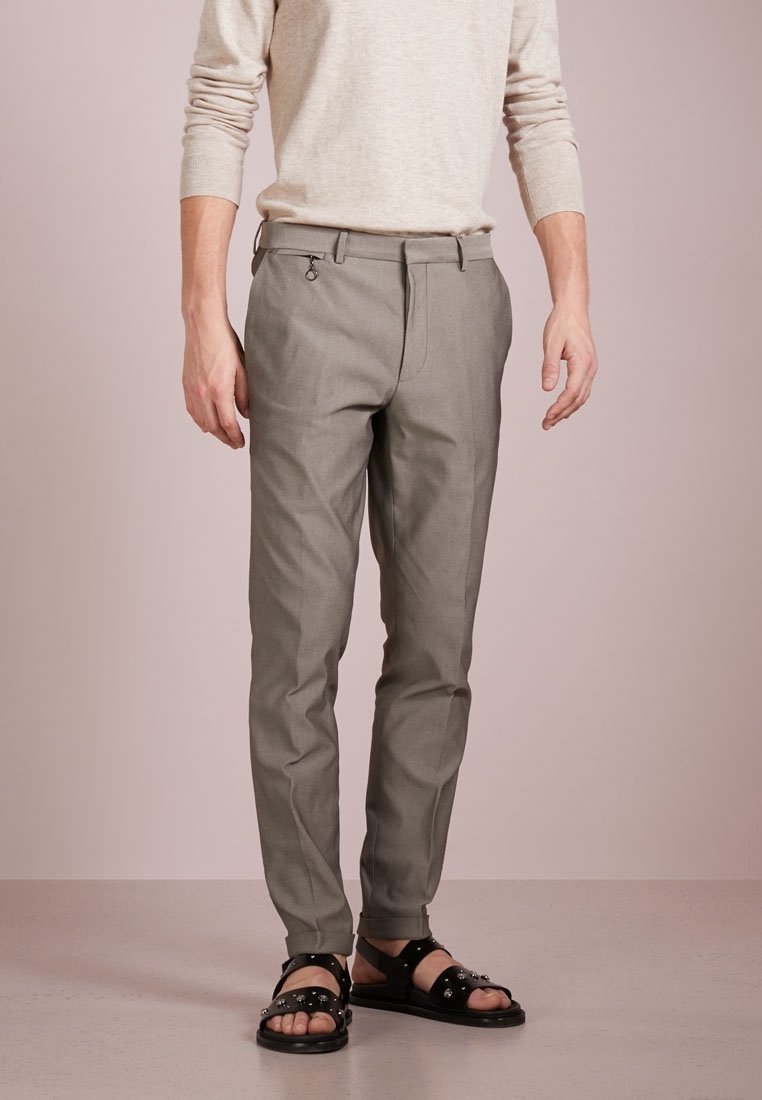 Homme portant un pull beige, un pantalon gris sur mesure avec passant de ceinture et détail anneau, et des sandales noires cloutées, debout sur un sol clair.