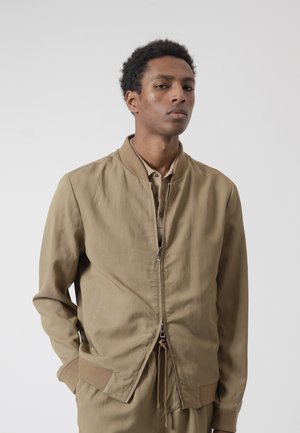 Joven con chaqueta bomber color beige sobre una camisa abotonada, con las manos en los bolsillos, posando frente a un fondo claro y uniforme.