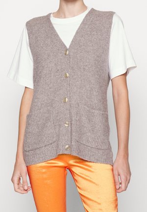 Personne portant un gilet beige tricoté boutonné avec poches par-dessus une chemise blanche à manches courtes et un pantalon orange brillant.