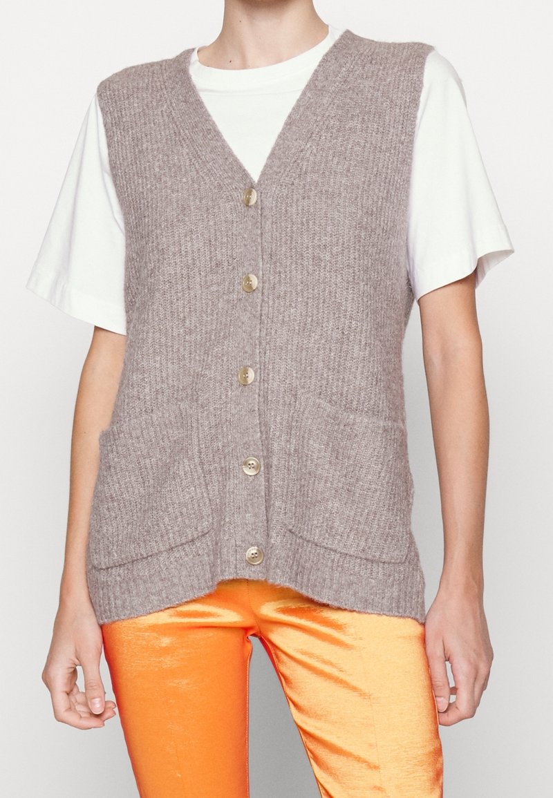 Personne portant un gilet beige tricoté boutonné avec poches par-dessus une chemise blanche à manches courtes et un pantalon orange brillant.