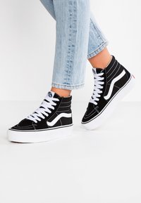 Persona che indossa jeans azzurri chiari e scarpe alte nere con lacci bianchi e suole bianche spesse, in piedi su una superficie bianca.