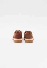 Chaussures en daim marron avec un bout arrondi, une languette à l'arrière et une semelle en caoutchouc blanc. Le design présente des coutures visibles et une surface texturée.