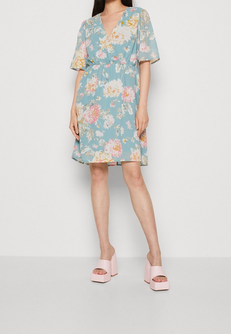 Femme portant une robe bleue à fleurs jusqu'aux genoux avec des manches courtes et des sandales compensées roses, debout devant un fond blanc uni.
