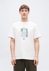 Άσπρο βαμβακερό t-shirt με άνετη γραμμή, που διαθέτει ένα πράσινο και γκρι γραφικό τύπωμα ενός αστροναύτη. Δεν είν αι εμφανής κάποια ιδιαίτερη υφή ή εξαρτήματα.