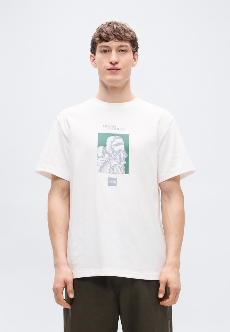 Άσπρο βαμβακερό t-shirt με άνετη γραμμή, που διαθέτει ένα πράσινο και γκρι γραφικό τύπωμα ενός αστροναύτη. Δεν είναι εμφανής κάποια ιδιαίτερη υφή ή εξαρτήματα.