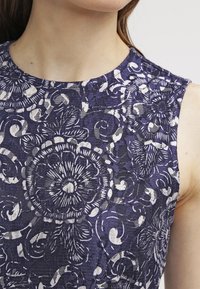 Gros plan sur une robe sans manches bleu foncé ornée de motifs complexes en dentelle florale blanche portée par une personne.