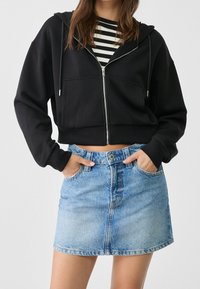 Sudadera con capucha negra recortada con bolsillos delanteros, combinada con una falda vaquera azul claro con cierre de botones y dos bolsillos laterales. Camiseta de rayas debajo.