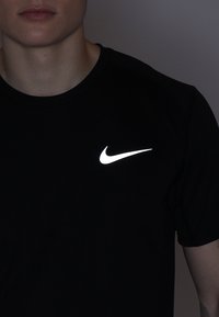 Czarna koszulka z krótkim rękawem wykonana z gładkiego materiału, z wyraźnym, odblaskowym białym logo Nike na lewej stronie klatki piersiowej.