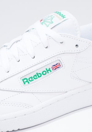 Gros plan d'une sneaker blanche Reebok avec logo vert, drapeau Union Jack, lacets blancs et détails en cuir cousu.