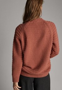 Sweter z fakturowanej koralowej wełny, z ribbowanym wykończeniem u dołu i przy mankietach, z raglanowymi rękawami i luźnym krojem, zaprezentowany z tyłu na neutralnym tle.