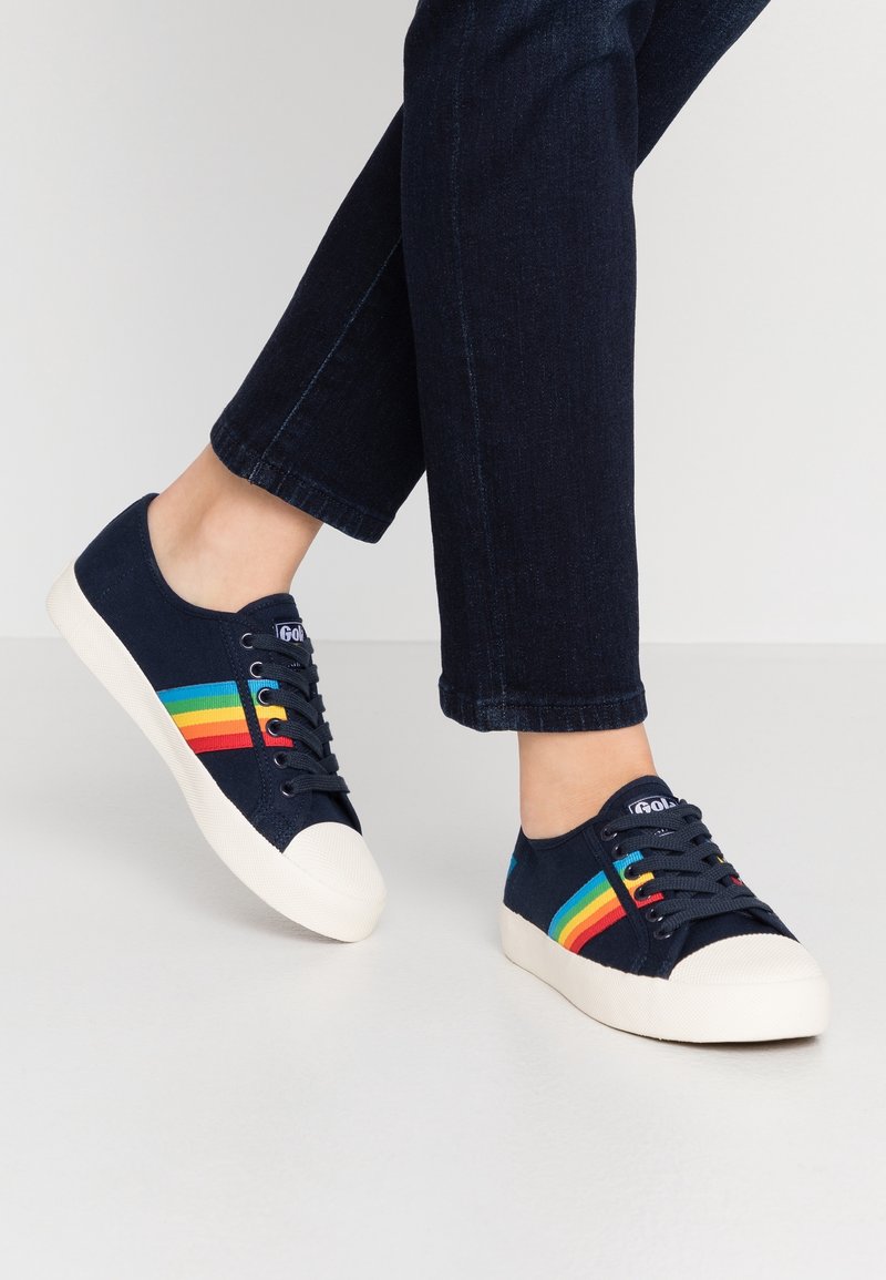 Personne portant des baskets bleu foncé avec des bandes arc-en-ciel et des semelles blanches, associées à un jean bleu foncé sur un fond uni.