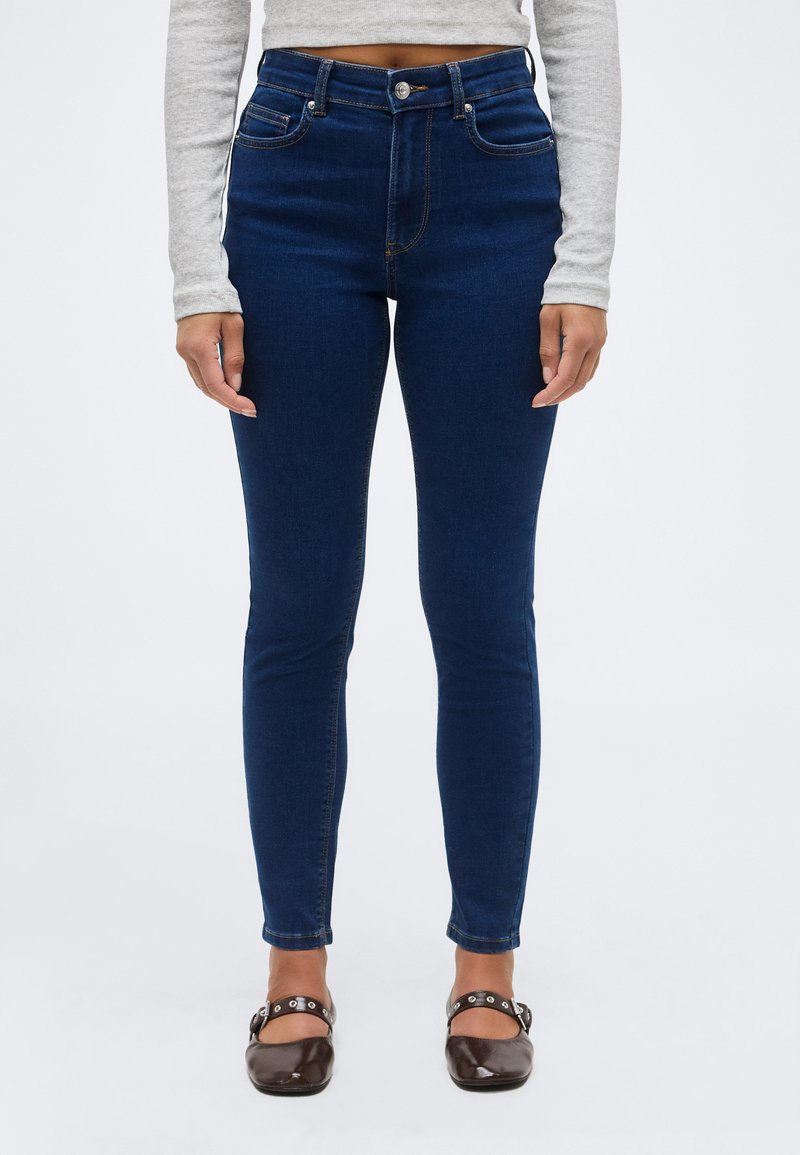 Γυναίκα που φοράει σκούρα μπλε στενά τζιν, γκρι μακρυμάνικη κοντή μπλούζα και σκούρα καφέ παπούτσια Mary Jane με λουράκια στον αστράγαλο, στέκεται σε λευκό φόντο.