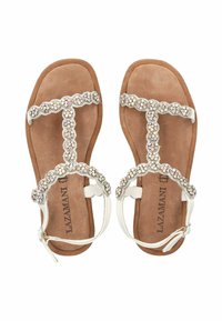 Weiße flache Sandalen mit T-Riemen-Design, verziert mit Strasssteinen, auf einer braunen Wildleder-Fußbett. Mit einem Riemen um den Knöchel.