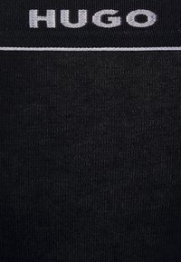 Leggings noirs avec une texture lisse, une taille élastique portant "HUGO" brodé en blanc et un détail en fine rayure blanche.