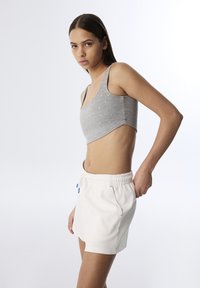 Joven mujer vistiendo un top corto gris con pequeños puntos blancos y pantalones cortos de cintura elástica blanca, posando contra un fondo claro y liso.