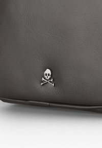 Bolso de cuero gris con un emblema de calavera y tibias metálico. La textura es lisa con líneas finas, y el diseño es minimalista.