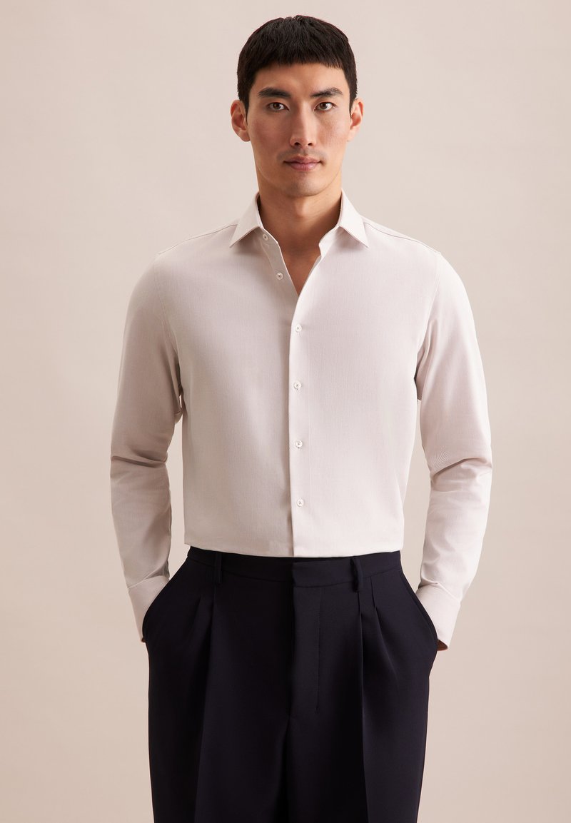 Lichtbeige langemouwshirt met een puntkraag en knopen aan de voorkant, gecombineerd met donkerblauwe broek. Stof lijkt glad en op maat gemaakt.