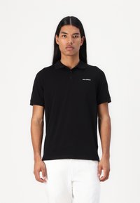 Černé polo tričko vyrobené z hladké tkaniny, s klasickým límečkem, krátkým rukávem a diskrétním logem na hrudi. Čistý a přiléhavý design.