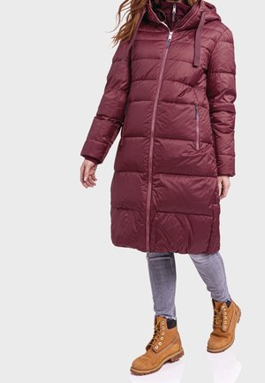 Bordeaux pufferjas met een capuchon, gequilt ontwerp, ritssluiting aan de voorkant en zijzakken, gecombineerd met tan veterschoenen en grijze jeans.