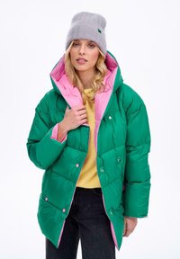 Zelená péřová bunda s růžovým podšíváním, s kapucí, knoflíky a prošívaným vzorem. Nositá přes žlutý top a s šedou čepicí.