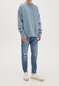 Persona che indossa un grande pullover azzurro, jeans blu strappati con buchi sulle ginocchia e sneakers bianche su uno sfondo semplice.