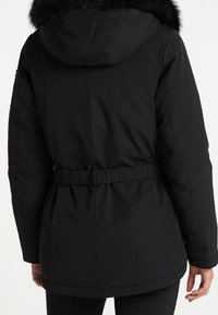 Parka negra con capucha que incluye un borde de piel sintética, cintura ceñida y acabado liso mate; diseñada para ofrecer calor y comodidad.