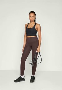 Fekete sportmelltartó, bordázott barna leggings, fekete edzőcipők, fehér bokazokni, ellenállás szalagot tart, semleges háttér.