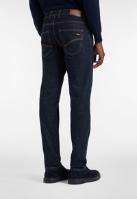 Moški nosi temno modre kavbojke z vidnimi zadnjimi žepe in rumenim šivom, v kombinaciji s temnomodrimi čevlji in temnomodro jopico.