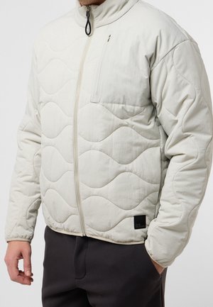 Mann trägt eine hellbeige gesteppte Jacke mit wellenförmiger Steppung, Reißverschluss vorne, Brusttasche und schwarzem Etikett über dem Saum.