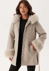 Cappotto in lana beige con cappuccio e polsini in pelliccia sintetica, dotato di una linea dritta, chiusura a bottoni e tasche laterali. Tessuto strutturato e caldo.