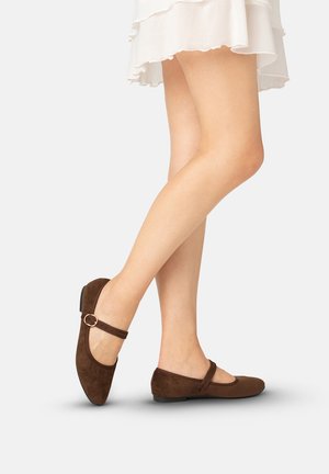 Nogi w brązowych zamszowych balerinach Mary Jane z paskami i biała falbaniasta spódnica na jasnym tle.