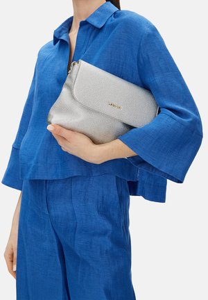 Mujer vestida con camisa y pantalones de lino azul, sosteniendo un bolso de mano plateado con textura y letras doradas a la altura de la cintura.