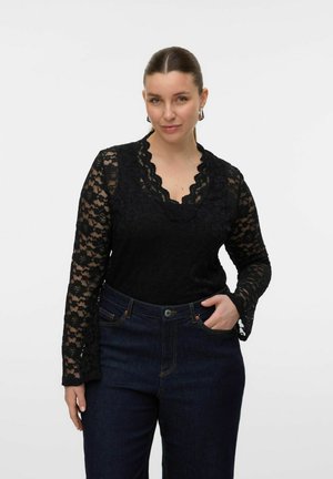 Schwarzes Spitzenoberteil mit langen Ärmeln, gewelltem Ausschnitt und aufwendigem Blumenmuster, kombiniert mit dunklen Jeans aus Denim mit Fronttaschen.