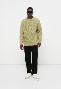 Olijfgroene trui met groot logopatroon, ruime pasvorm, lange mouwen, gemaakt van textuurgebreid materiaal, gecombineerd met zwarte broek en witte sneakers.