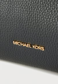 Sort tekstureret læderpung med en guldfarvet "MICHAEL KORS"-logo foran. Overfladen har et diskret kornmønster.