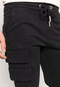 Černé cargo kalhoty vyrobené z bavlny, s páskem na stažení, bočními kapsami a dvěma velkými nášivkami na stehnech.