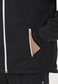 Sort jakke med hvid lynlås, to sidelommer og en glat overflade. Har en tætsiddende pasform og elastiske håndled.