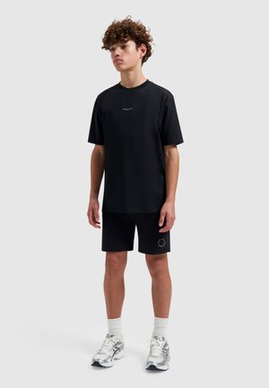Tiener met krullend haar, gekleed in een zwart T-shirt en korte broek, witte sportsokken en zilveren sportschoenen, staand tegen een effen lichte achtergrond.