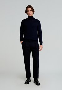 Schwarzer Rollkragenpullover aus weichem Material, kombiniert mit dunklen, maßgeschneiderten Hosen. Das Model steht selbstbewusst mit den Händen in den Taschen vor einem minimalistischen Hintergrund.