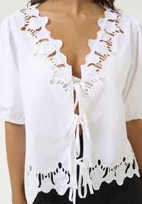 Blusa bianca in cotone con scollatura a V profonda, caratterizzata da ritagli decorativi e un bordo in pizzo. I legami frontali aggiungono dettagli, e le maniche corte a sbuffo completano il design.