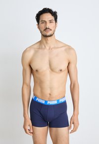 Sötétkék boxeralsó, kék márkázott gumírozással. Puha, sima anyag, szoros illeszkedés és minimalista dizájn. A modell frontálisan áll.